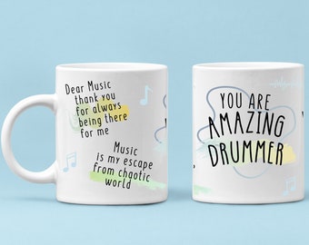 Op de afbeelding: Een witte keramische mok met een blauw en geel aquarelontwerp. De mok heeft de tekst "Dear Music thank you for always being there for me Music is my escape from chaotic world" aan de ene kant en "YOU ARE AMAZING DRUMMER" aan de andere kant.