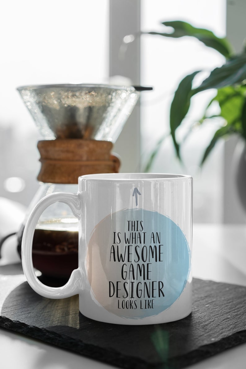 May include: White ceramic coffee mug with a blue and peach watercolor circle design. The text on the mug reads "This is what an awesome game designer looks like".