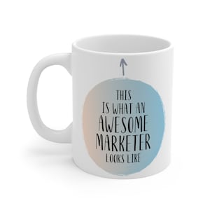May include: White ceramic coffee mug with a blue and orange circle design. The circle has the text "This is what an awesome marketer looks like" inside. An arrow points up to the circle.