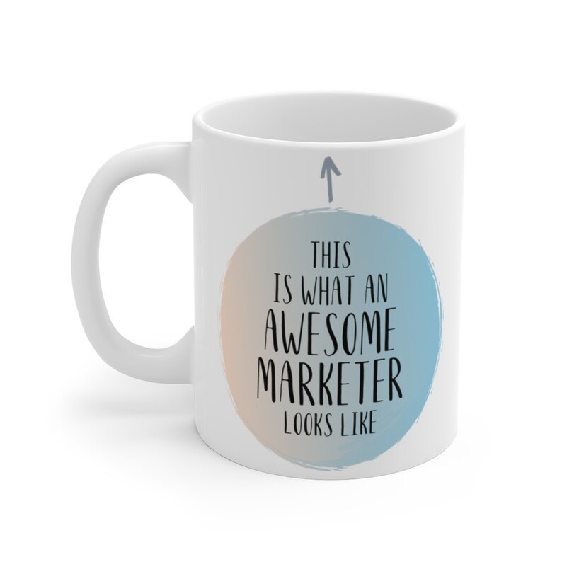 May include: White ceramic coffee mug with a blue and orange circle design. The circle has the text "This is what an awesome marketer looks like" inside. An arrow points up to the circle.