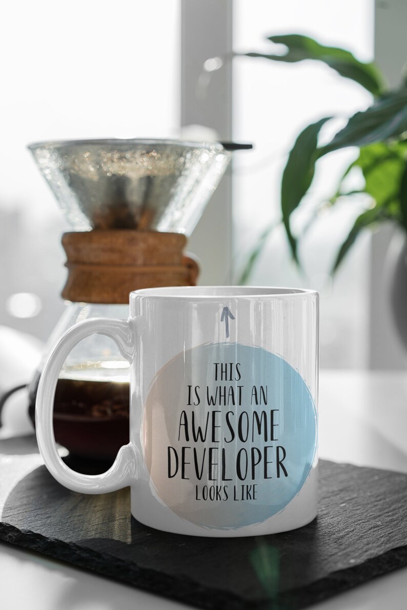 Pode incluir: Caneca de caf&eacute; de cer&acirc;mica branca com um design circular azul e p&ecirc;ssego. A caneca tem uma al&ccedil;a preta e o texto "This is what an awesome developer looks like" impresso no c&iacute;rculo.