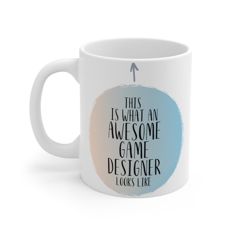 May include: White ceramic coffee mug with a blue and peach watercolor circle design. The text inside the circle reads "This is what an awesome game designer looks like". An arrow points up to the circle.