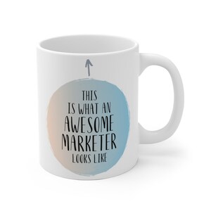 May include: White ceramic coffee mug with a blue and orange watercolor circle design. The circle has the text "This is what an awesome marketer looks like" in black.