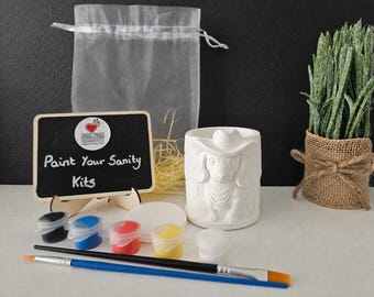 May include: A craft kit featuring a white ceramic dachshund figurine, a small chalkboard sign with the text Paint Your Sanity Kits, five small paint pots in black, blue, red, yellow, and white, two paintbrushes, and a sheer drawstring bag.