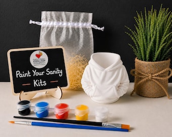 May include: A DIY painting kit featuring a white ceramic hoodie-shaped planter, a small chalkboard sign with the text "Paint Your Sanity Kits", a sheer drawstring bag with filler, five small paint pots in various colors, and two paintbrushes.