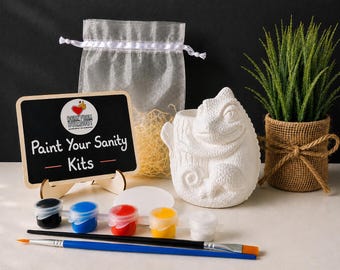 May include: A paint-your-own ceramic chameleon figurine kit including a small chalkboard sign with the text Paint Your Sanity Kits, a sheer white drawstring bag, five small paint pots in black, blue, red, yellow, and white, and two paintbrushes.