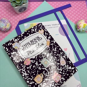 May include: A composition notebook with a black and white speckled design and the text "THIS BOOK BELONGS TO Mrs. Silva". The notebook has colorful stickers of a globe, a pencil, and a coffee mug. A purple T-square and a small succulent are also visible.