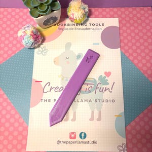 May include: A purple bookbinding tool rests on a card with the text "Creating is fun!" and a llama illustration. The card is on a pink and blue patterned surface, with a small succulent and colorful yarn pom-poms.