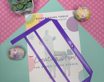 May include: A purple bookbinding tool with a rectangular frame and diagonal supports, resting on a card with the text "Creating is fun!" and a llama illustration. The background features a succulent, pom-poms, and a pink and teal color scheme.