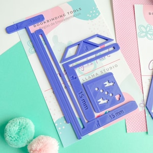 May include: Purple plastic bookbinding tools with measurements in millimeters. The tools include a ruler, a corner creaser, and a spine stitching tool. The tools are on a white background with pink and white polka dots.