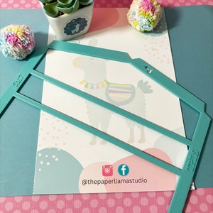 May include: A turquoise blue plastic stencil shaped like a house with two horizontal lines and the words "5x7" and "4x6" printed on it. The stencil is on top of a white card with a cartoon llama illustration and pink polka dots. The card has the text "@thepaperllamastudio" printed on it.