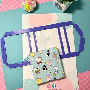 May include: A blue plastic template with the text "3x2.5" printed on it. The template has three rectangular cutouts. A small blue and white fabric pouch with a Hello Kitty character design is placed on top of the template. The pouch has a blue string attached to it. The background is a pink and blue polka dot pattern.