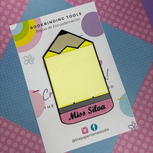 May include: A yellow pencil shaped bookmark with a pink eraser. The eraser has the text "Miss Silva" written on it. The bookmark is on a white background with a grid pattern and the text "BOOKBINDING TOOLS Reglas de Encuaderancion" at the top.