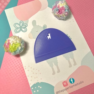 May include: A purple llama shaped die cut with a white llama silhouette inside. The die cut is on a white card with a pink and blue background. The card also has a white flower and a white llama with a pink nose and blue eyes. The card has the text "@thepaperllamastudio" and social media icons.