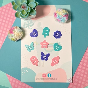May include: A set of 10 colorful die-cut shapes, including a heart, star, butterfly, and ice cream cone. The shapes are made of paper and have a light blue, pink, and purple color scheme. The shapes are arranged on a white background with a pink and white polka dot pattern.