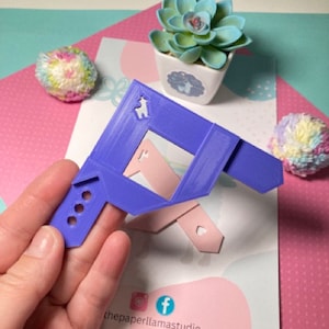 May include: A purple 3D-printed frame with a llama logo, held in a hand. A light pink piece is attached to the frame. A small succulent in a white pot and two colourful pom-poms are in the background.