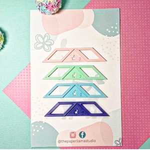 May include: Set of five colorful plastic triangle rulers with a small llama graphic on each. The rulers are stacked on top of each other, with the pink ruler on top and the blue ruler on the bottom. The rulers are on a white background with a pink and green abstract design.