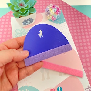 May include: Purple felt craft stencil with a llama cutout. The stencil has a purple felt border and a pink felt backing. The stencil is held in a hand.