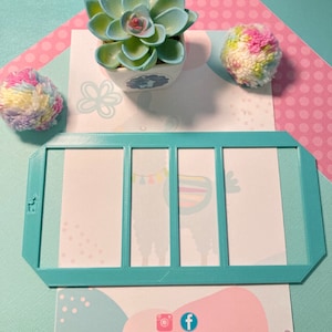 May include: A turquoise blue plastic template with three rectangular cutouts. The template is on a white background with pink polka dots and a small succulent plant in a white pot.