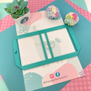 May include: Teal plastic stencil with a 3x2.5 inch rectangle cut out. The stencil is designed for crafting and features a llama illustration. The stencil is on a pink and blue background with white polka dots.