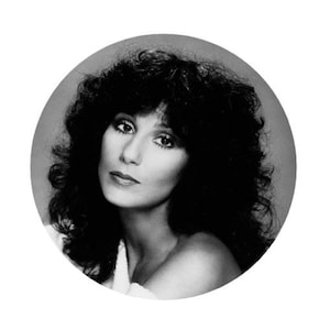 May include: Black and white portrait of a woman with long, dark, curly hair. She is wearing a white top and looking directly at the camera.
