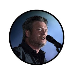 May include: A close-up of a man singing into a microphone. He is wearing a black shirt and has a beard. The background is a blurred blue.