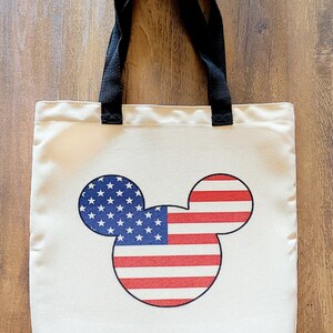 May include: A white canvas tote bag with black handles featuring a Mickey Mouse silhouette with the American flag design.