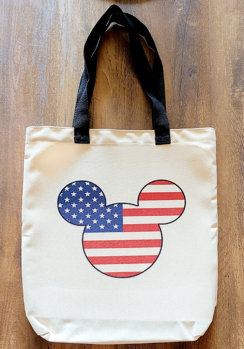 May include: A white canvas tote bag with black handles featuring a Mickey Mouse silhouette with the American flag design.