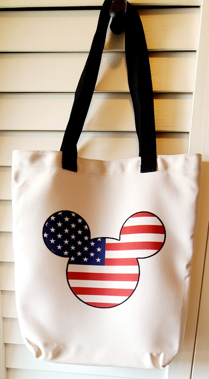 May include: A white canvas tote bag with black straps featuring a Mickey Mouse silhouette design. The silhouette is filled with the American flag pattern.