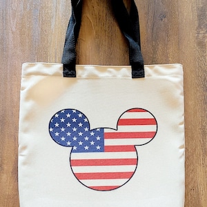 May include: A white canvas tote bag with black handles featuring a Mickey Mouse silhouette with the American flag design.