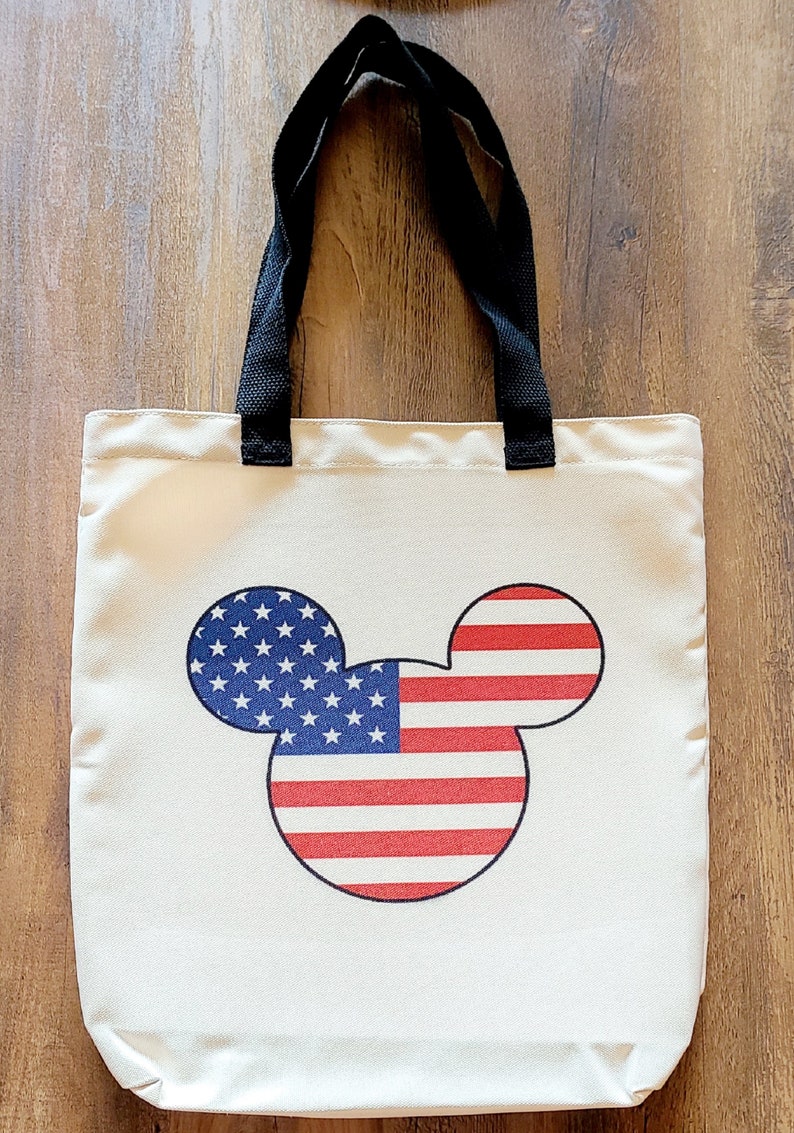May include: A white canvas tote bag with black handles featuring a Mickey Mouse silhouette with the American flag design.