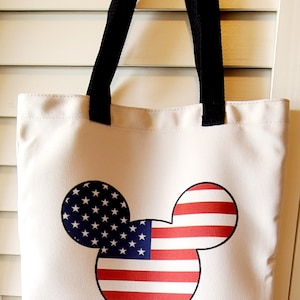 May include: A white canvas tote bag with black straps featuring a Mickey Mouse silhouette design. The silhouette is filled with the American flag pattern.