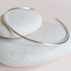 May include: A silver cuff bracelet with a hammered texture. The open-ended bracelet is displayed on a white stone against a soft pink background. The bracelet is a simple, elegant piece of jewellery.