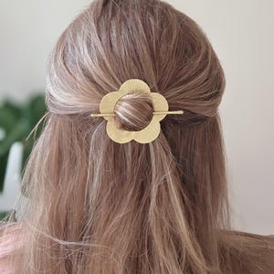 May include: A gold-coloured flower-shaped hair clip is secured in the back of the hair. The hair clip has a textured surface and a bar through the centre of the flower. The hair is light brown and wavy.