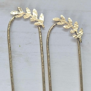 May include: Two gold hair pins with a leaf design at the top. The pins are long and thin, and the leaves are hammered for a textured look.