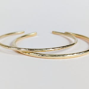 Peut inclure: Deux bracelets ouverts dorés sont présentés sur un fond blanc. Les bracelets ont une texture martelée et un design simple et élégant. Les bijoux sont probablement en métal.