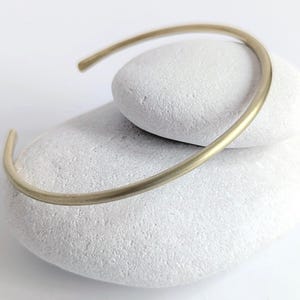 May include: A minimalist gold-coloured cuff bracelet. The open-ended bracelet has a smooth, rounded design. The jewellery is displayed on two white, smooth stones, creating a simple, elegant aesthetic. The bracelet is a versatile accessory.