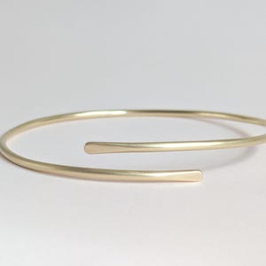May include: A gold-colored open cuff bracelet. The bracelet has a simple, minimalist design with a smooth, matte finish. The ends of the bracelet are slightly flattened. The bracelet is a jewelry item.