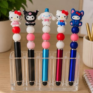 May include: Five pens with character toppers in a clear acrylic holder. Each pen features a unique silicone character, including Hello Kitty and Kuromi, atop a stack of colorful beads. The pen barrels are black, blue, and red with metallic finishes.