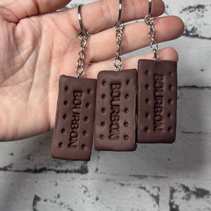 May include: Three rectangular brown keychains shaped like Bourbon biscuits, each featuring the embossed word BOURBON and small circular indentations, attached to silver-coloured metal split rings by short silver-coloured metal link chains.