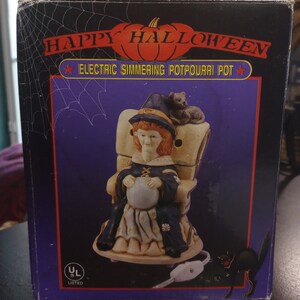 May include: A box for an electric simmering potpourri pot. The box features a witch sitting in a chair with a black cat on her shoulder. The box is purple with a white spider web design. The text on the box reads "Happy Halloween Electric Simmering Potpourri Pot".