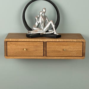 May include: A floating wooden shelf with two drawers featuring small round knobs. Positioned on top of the shelf is a decorative sculpture of two silver figures sitting within a black circular frame, creating a balanced and structured display.