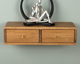 May include: A floating wooden shelf with two drawers featuring small round knobs. Positioned on top of the shelf is a decorative sculpture of two silver figures sitting within a black circular frame, creating a balanced and structured display.
