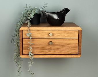 May include: A floating wooden nightstand with two drawers featuring round brass-colored knobs. A black bird-shaped ceramic figurine and a trailing green plant in a black pot are placed on top of the light brown wood grain surface.