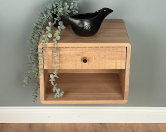May include: A light brown wooden floating nightstand with a single drawer featuring a vertical slat design and a small round knob. A black bird-shaped ceramic decorative object and a trailing green plant are placed on top of the nightstand.