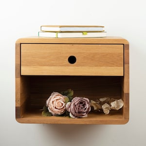 May include: A light brown, wooden floating bedside table with a drawer and open shelf. Two notebooks rest on top. The drawer has a circular handle. Artificial flowers in pink and purple are in the open shelf.