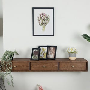 Puede incluir: Una estanter&iacute;a flotante de color marr&oacute;n oscuro con tres cajones y pomos de lat&oacute;n. Encima de la estanter&iacute;a hay tres cuadros enmarcados y una impresi&oacute;n bot&aacute;nica enmarcada de una alcachofa. Se ven una peque&ntilde;a planta en maceta y una l&aacute;mpara.