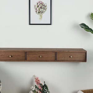 Puede incluir: Una estanter&iacute;a flotante de madera con tres cajones. La estanter&iacute;a es de color marr&oacute;n c&aacute;lido y tiene pomos de lat&oacute;n. Los cajones est&aacute;n espaciados uniformemente. La estanter&iacute;a est&aacute; montada en una pared blanca, con una impresi&oacute;n bot&aacute;nica enmarcada encima.