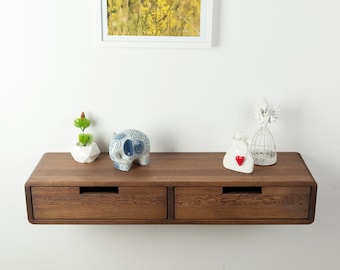May include: A floating wall-mounted wooden shelf featuring two drawers with horizontal cut-out handles. The shelf has a dark brown wood grain finish and is decorated with a small green plant, a blue and white patterned elephant, and two white ornaments.