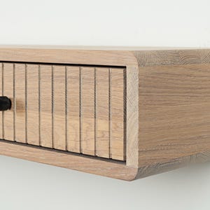 May include: A light brown wooden floating wall shelf featuring a single drawer with a vertical slatted front design and a small, round, black knob. The shelf has rounded edges and a visible natural wood grain texture throughout the entire structure.